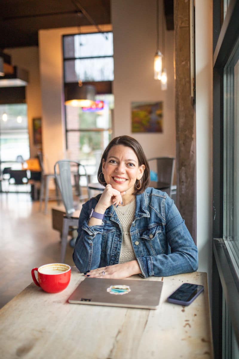 Photo of Laura Jawad sitting at a table in a coffee shop, chin resting on her closed hand, closed laptop on the table in front of her.