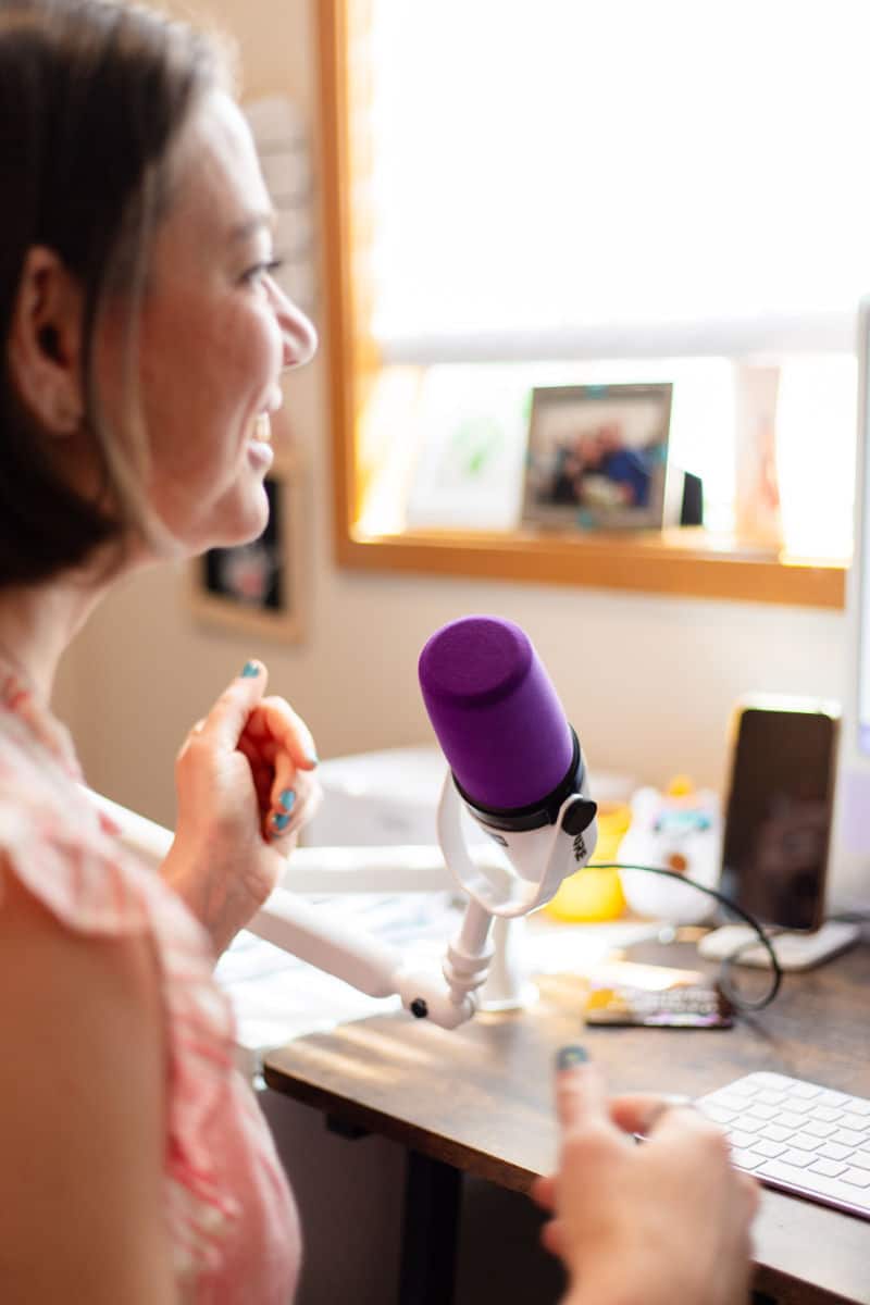 Laura Jawad smiling and speaking into a purple microphone at her home office desk, with a keyboard and phone visible in the background.